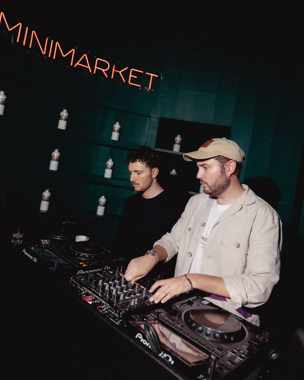 Two DJs on the decks under the Minimarket neon sign