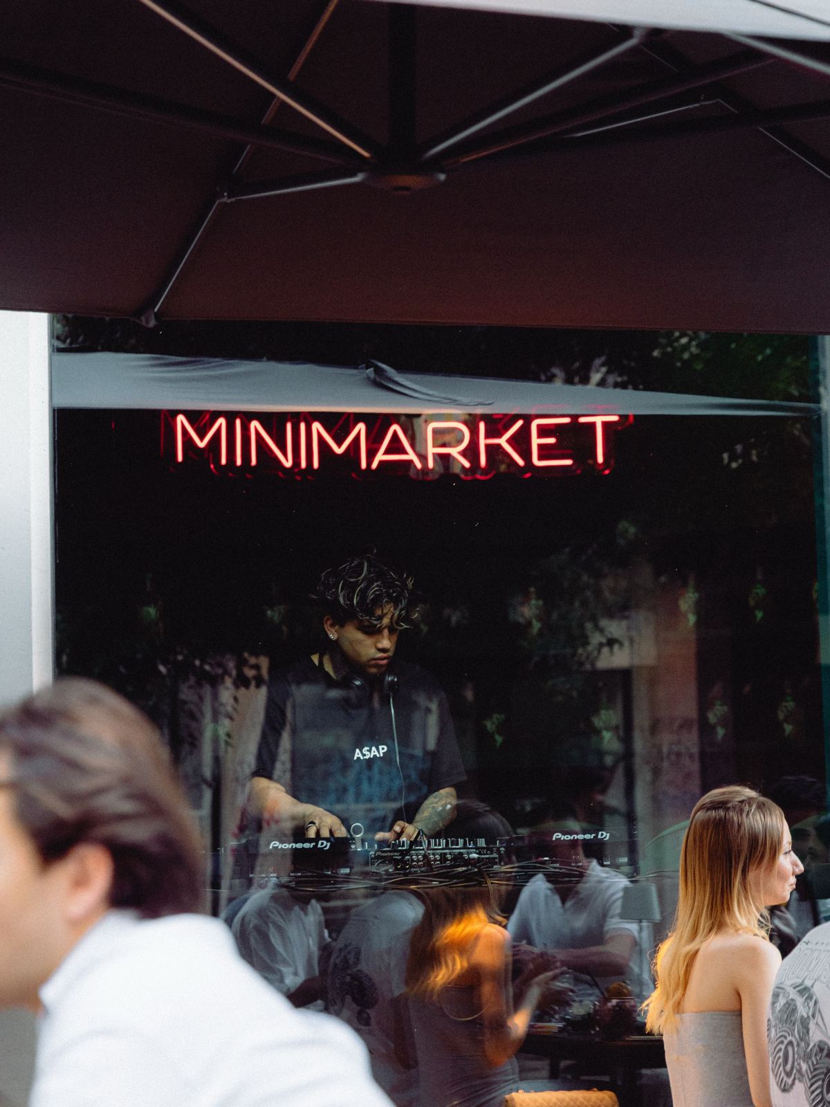 The entrance of Minimarket at night, neon sign on, people chatting out front